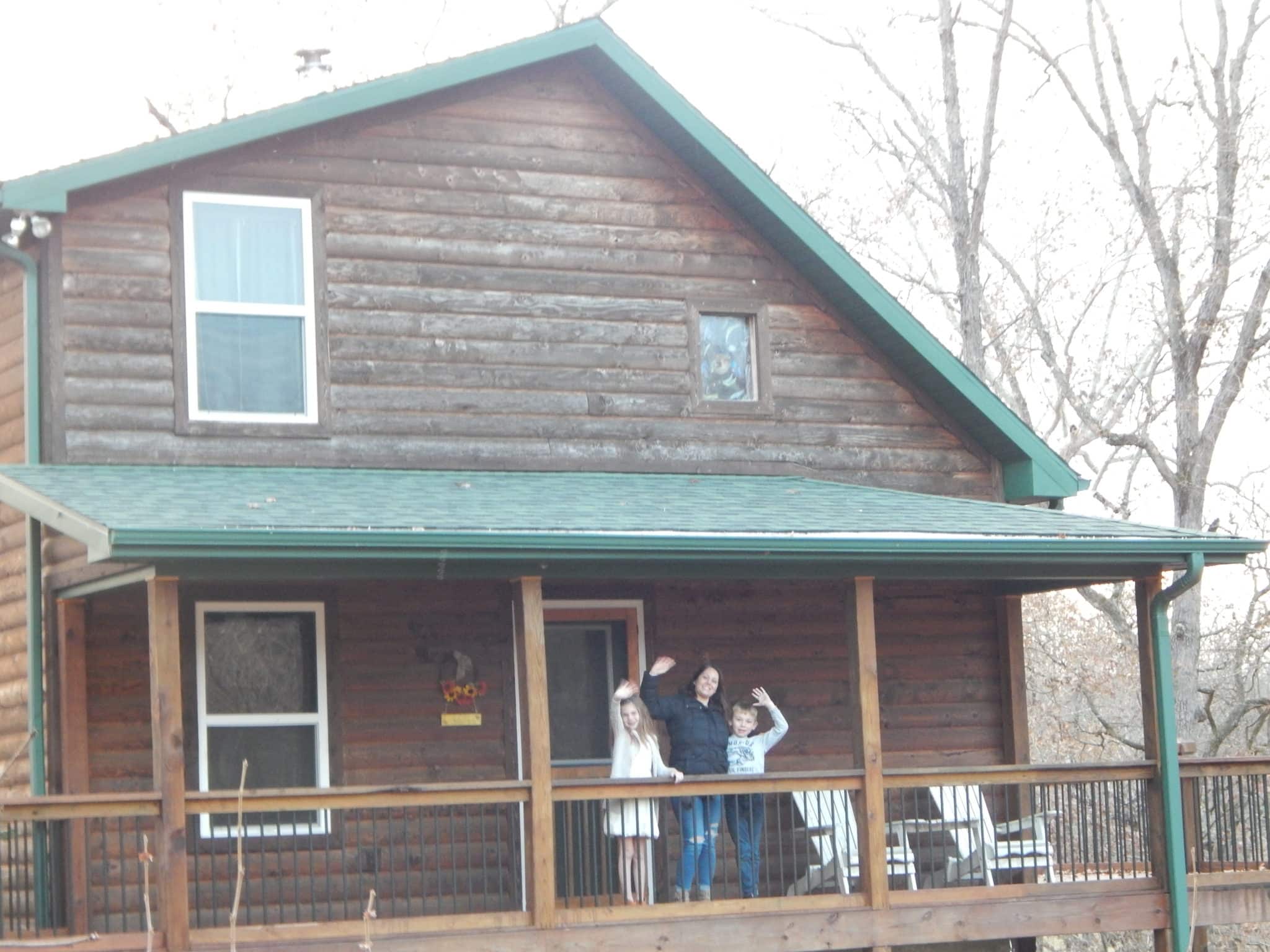 Woman and two children waving from the front porch of cabin.