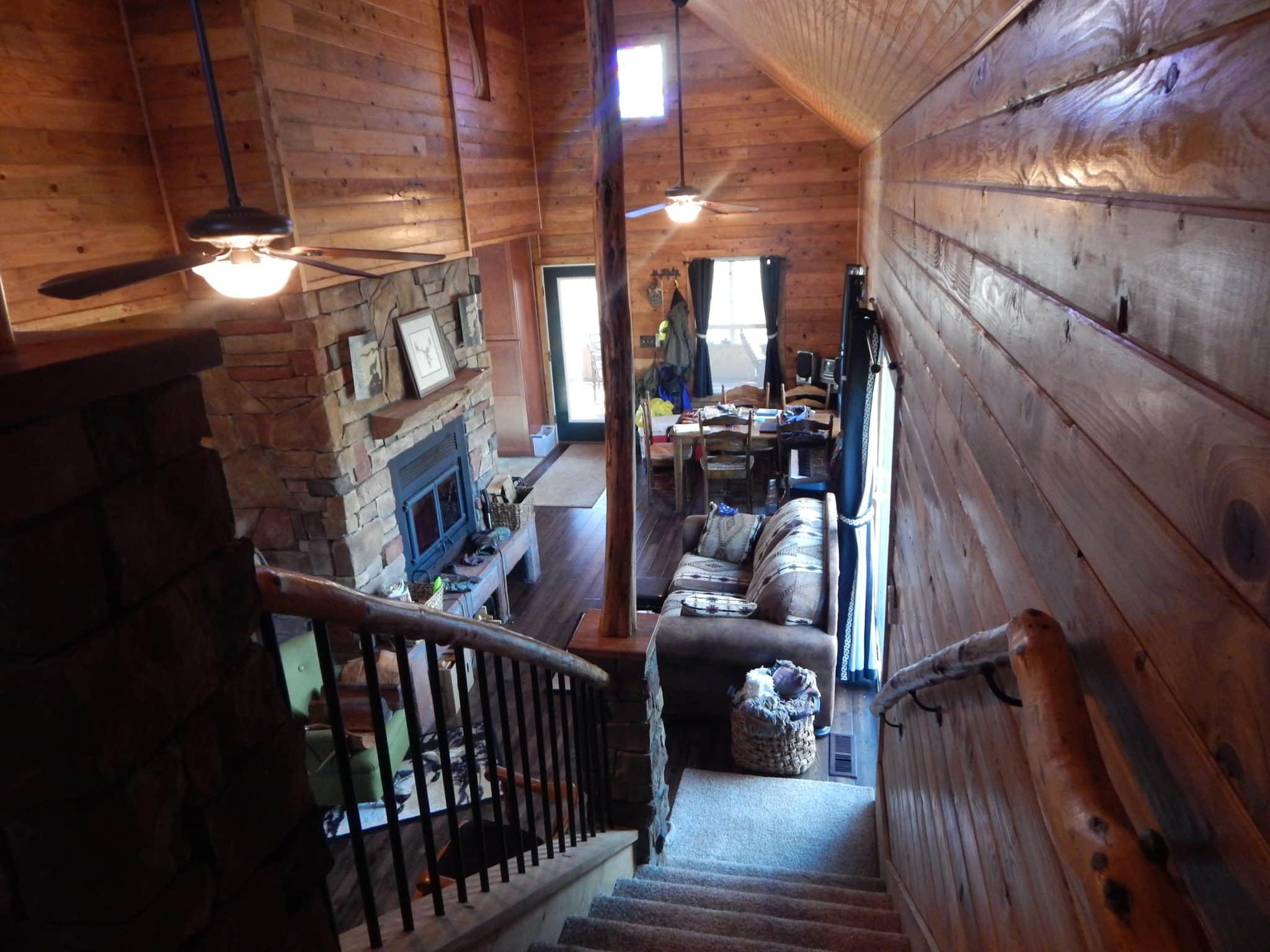 Looking down the stairs to the cabin floor showing the fireplace, couch, fan lights and back porch glass door.