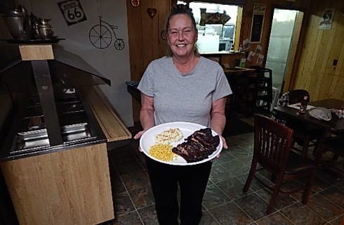 Lady with gray shirt standing in restaurant holding a plate full of barbequed ribs, corn, and mashed potatoes