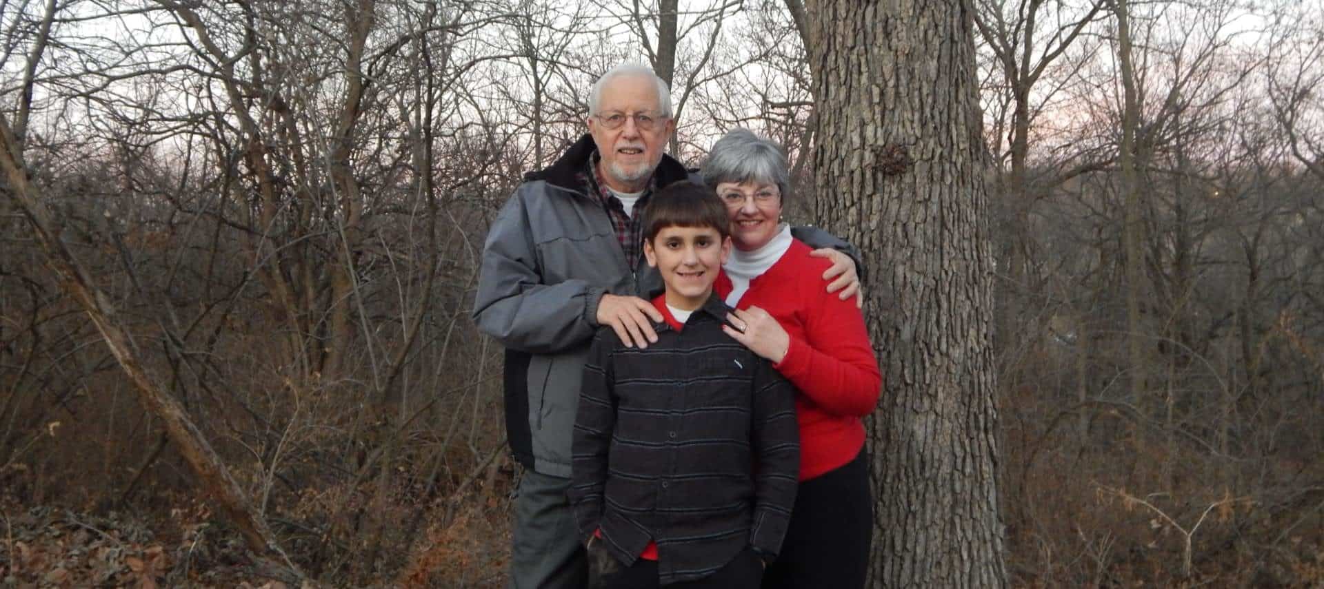 Man with gray jacket standing with lady with red sweater and boy with dark gray button up shirt near trees and bushes with no leaves